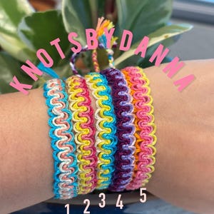 May include: A collection of five colorful handmade friendship bracelets. Each bracelet features a unique combination of vibrant colors, including blue, pink, yellow, purple, and orange. The bracelets are labeled with numbers 1 through 5.