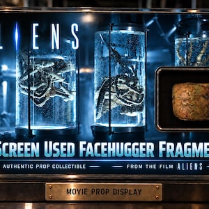 Może przedstawiać: Prostokątna gablota z napisem ALIENS oraz A SCREEN USED FACEHUGGER FRAGMENT. Wewnątrz znajdują się dwa cylindryczne szklane pojemniki z okazami organicznymi oraz mały, teksturowany fragment w kolorze opalenizny na czarnym tle.