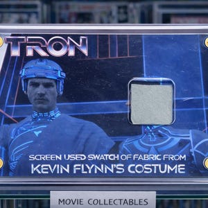 Könnte beinhalten: Ein gerahmtes Filmsammlerstück mit dem TRON-Logo und einem Stoffmuster aus Kevin Flynns Kostüm. Das Bild hat einen Blauton, ein Porträt der Figur und den Text "SCREEN USED SWATCH OF FABRIC FROM KEVIN FLYNN'S COSTUME."