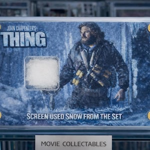 Op de afbeelding: Een ingelijst filmverzamelobject van John Carpenter's "The Thing". De afbeelding toont een man in een winterjas en de tekst "SCREEN USED SNOW FROM THE SET". De lijst heeft goudkleurige accenten en een wit vierkant sneeuw.