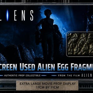 May include: A framed movie prop display from the film "Aliens." The display features the film's title, a silhouette of characters, and a fragment of an alien egg. The display measures 17cm by 11cm.