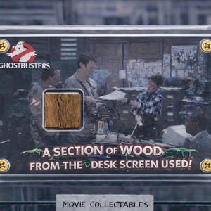 Op de afbeelding: Een Ghostbusters-filmverzameling met een stuk hout van het bureauscherm. De display bevat het Ghostbusters-logo en de tekst "A SECTION OF WOOD FROM THE DESK SCREEN USED!" in het groen. Het verzamelobject zit in een doorzichtige display.