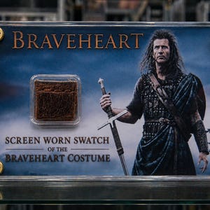 Op de afbeelding: Een ingelijste display met een op het scherm gedragen staal van het Braveheart-kostuum. De display bevat een klein, vierkant stuk bruine stof en een afbeelding van de hoofdpersoon van de film. De tekst "BRAVEHEART" staat bovenaan.