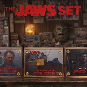 Op de afbeelding: De Jaws Set-display met drie ingelijste items met filmmemorabilia. Elk frame heeft een gouden rand en messing schroeven. De tekst "JAWS" staat bovenaan elk frame. De frames bevatten een stuk van Matt Hoopers shirt, een dekgedeelte en Shark's Tooth.