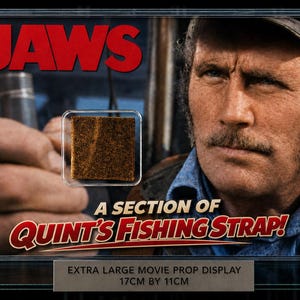 Op de afbeelding: Een ingelijste filmrekwisieten display met het woord "JAWS" in het rood, een portret van een acteur en een gedeelte van Quint's visriem. De display is 17 cm bij 11 cm.