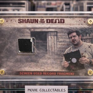 May include: A framed movie collectible from "Shaun of the Dead" featuring a screen-used record fragment. The image includes the movie title and a photo of an actor holding a record. The collectible is encased in a clear acrylic case.