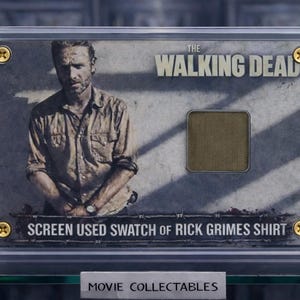 May include: A display case containing a screen-used fabric swatch from Rick Grimes' shirt, as seen in The Walking Dead. The image includes a portrait of the character and the show's logo. The case is secured with gold-coloured screws.