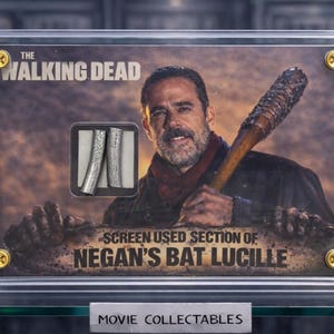 Op de afbeelding: Een ingelijst verzamelobject van "The Walking Dead" met een sectie van Negans knuppel, Lucille. De afbeelding bevat een portret van het personage en de tekst "SCREEN USED SECTION OF NEGAN'S BAT LUCILLE". Het frame is bevestigd met goudkleurige schroeven.