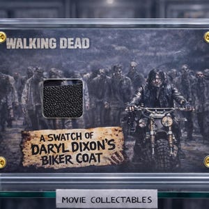 Op de afbeelding: Een ingelijst verzamelobject met een stukje van Daryl Dixon's bikerjas uit The Walking Dead. De afbeelding toont Daryl op een motor met zombies op de achtergrond. De lijst heeft gouden accenten en de tekst "Movie Collectables".