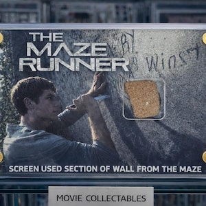 Op de afbeelding: Een ingelijst filmverzamelobject van "The Maze Runner" met een gedeelte van een muur. De afbeelding toont een man die aan de muur werkt met de filmtitel bovenaan. De lijst heeft goudkleurige accenten en de tekst "SCREEN USED SECTION OF WALL FROM THE MAZE".