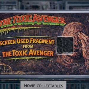 Op de afbeelding: Een ingelijst verzamelobject uit de film "The Toxic Avenger". De afbeelding toont de filmtitel in groen en rood, met een verweerde afbeelding van een personage. Het frame bevat een op het scherm gebruikt fragment en is vastgezet met goudkleurige schroeven.
