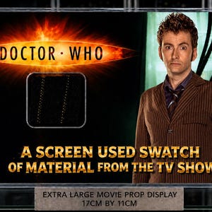 Op de afbeelding: Een ingelijste display met het "Doctor Who"-logo en een schermstaal. De display bevat de tekst "A SCREEN USED SWATCH OF MATERIAL FROM THE TV SHOW" en meet 17 cm bij 11 cm.