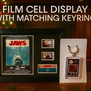 a framed poster of the movie "Jaws" with a keyring, a small keychain, and a framed picture of a shark. The background is blurred, but it appears to be a festive setting with Christmas lights.