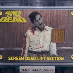 Op de afbeelding: Een ingelijst film verzamelobject van "Dawn of the Dead" met een zombie-afbeelding met bloedvlekken. De tekst "DAWN OF THE DEAD" is geel. De tekst "SCREEN USED LIFT SECTION" staat eronder. Het verzamelobject zit in een doorzichtige behuizing.