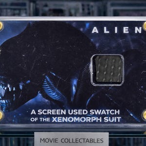Può includere: Un oggetto da collezione incorniciato con l'Alien Xenomorph. L'immagine mostra la testa della creatura e la parola "ALIEN" in bianco. Include un campione usato sullo schermo del costume Xenomorph. La cornice è fissata con viti color oro.