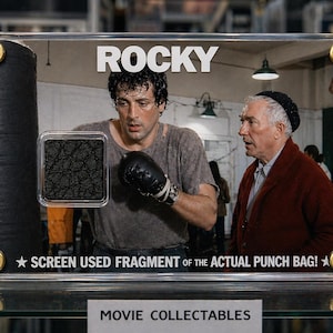 Puede incluir: Un objeto de colección de películas enmarcado con la película "Rocky". La imagen muestra un fragmento del saco de boxeo real, con Sylvester Stallone. El texto "ROCKY" está en la parte superior.