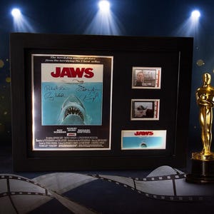 a framed movie poster for the film "Jaws", along with a golden Oscar statuette and film strips, all set against a dark background with spotlights shining down.