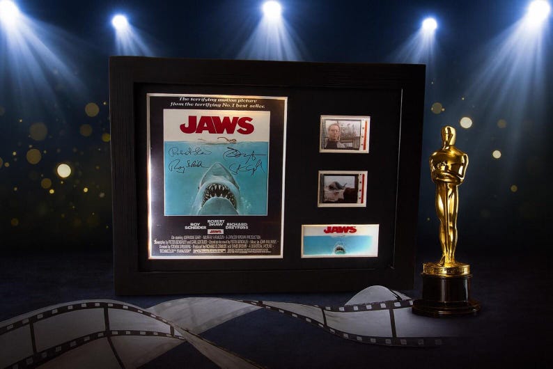 a framed movie poster for the film "Jaws", along with a golden Oscar statuette and film strips, all set against a dark background with spotlights shining down.