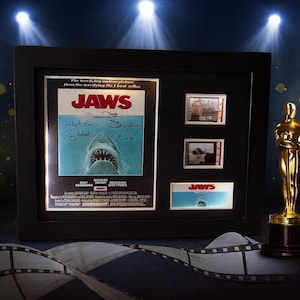 a framed movie poster for the film "Jaws", along with a golden Oscar statuette and film strips, all set against a dark background with spotlights shining down.
