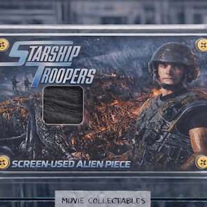 Pode incluir: Um item de colecionador do filme Starship Troopers, em uma vitrine de acrílico transparente. O cartão apresenta o título do filme, um soldado e uma peça alienígena usada na tela. O item de colecionador é fixado com parafusos dourados.