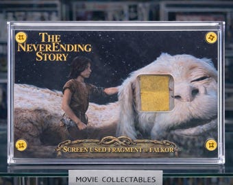 May include: A framed collectible featuring artwork from "The NeverEnding Story." The image shows a scene with a boy and Falkor, the Luckdragon, with a gold-colored screen-used fragment. The text "SCREEN USED FRAGMENT OF FALKOR" is visible.