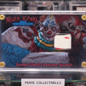 Op de afbeelding: Een ingelijst verzamelobject met de "Killer Klowns from Outer Space". De afbeelding toont drie clowngezichten met rode, witte en blauwe accenten. Inclusief een stofstaal van het kostuum dat op het scherm is gebruikt. De tekst "Screen Used Costume Swatch" is zichtbaar.