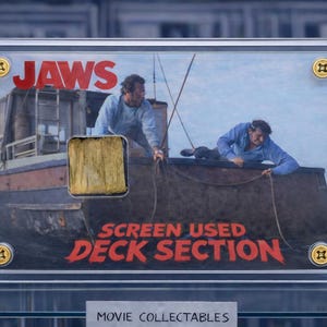 Op de afbeelding: Een ingelijst filmverzamelobject met het "Jaws"-filmlogo en een deel van het dek. De afbeelding toont twee mannen op een boot. De tekst "SCREEN USED DECK SECTION" is rood.