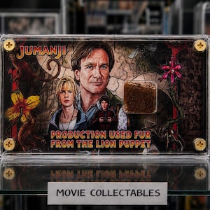 Op de afbeelding: Een ingelijste Jumanji-filmverzameling met afbeeldingen van acteurs en een stukje vacht van de leeuwenpop. De tekst luidt "PRODUCTION USED FUR FROM THE LION PUPPET".
