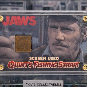 May include: A framed movie collectible from the film "Jaws." The image features a portrait of an actor, the film's title in red, and the text "SCREEN USED QUINT'S FISHING STRAP!" A small, square piece of the prop is visible.