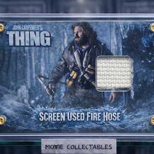 Puede incluir: Un objeto de colección de películas enmarcado de "The Thing" de John Carpenter. La obra de arte presenta a un hombre en un entorno nevado, con el título de la película y el texto "SCREEN USED FIRE HOSE". El marco está asegurado con tornillos dorados, y la parte inferior dice "MOVIE COLLECTABLES."