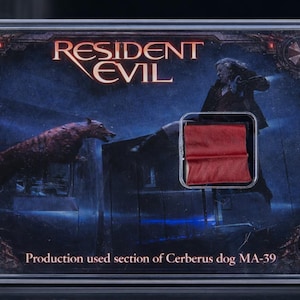 Pode incluir: Um item colecionável de Resident Evil emoldurado com uma ilustração escura de um cão Cerberus e uma figura. O texto "RESIDENT EVIL" está no topo, com "Production used section of Cerberus dog MA-39" na parte inferior. Um pedaço de tecido vermelho é visível.