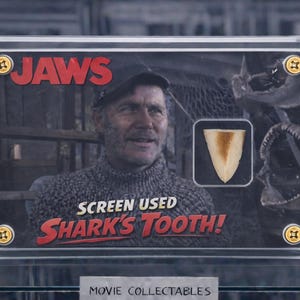 Op de afbeelding: Een ingelijst filmverzamelobject uit de film "Jaws". De afbeelding toont een man, een haaientand en de tekst "SCREEN USED Shark's Tooth!". Het "Jaws"-logo is rood. Het verzamelobject wordt in een doorzichtige behuizing getoond.
