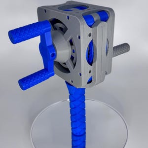 May include: A 3D-printed mechanical device with a gray, blue, and white color scheme. The device features a cube-shaped body with visible gears and a blue handle. The base is a blue, textured column. The device is displayed on a reflective surface.