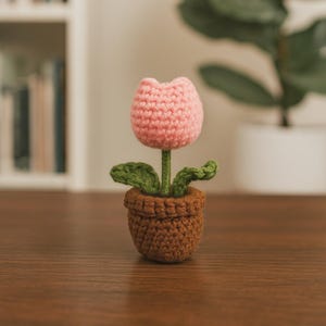 以下が含まれることがあります： 手作りのかぎ針編みのチューリップ、茶色の鉢植え。 ピンクの花には緑の葉と緑の茎があります。 鉢は濃い茶色です。 花は木製の表面にあります。