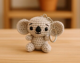 May include: A crocheted koala keychain in light gray yarn. The koala has large, round ears, a black nose, and small black eyes. A silver-colored chain and keyring are attached to the top. The koala is sitting on a wooden surface.