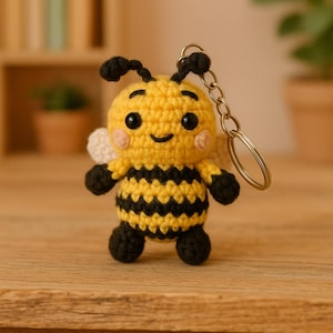 May include: A crocheted bee keychain with black and yellow stripes, black eyes, and a smiling mouth. The bee has white wings and is attached to a silver key ring.