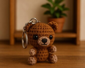 May include: A brown crocheted teddy bear keychain with black eyes and a light brown snout. The bear has a silver key ring attached to its head. The bear is sitting on a wooden surface.