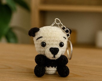 May include: A handmade crochet panda bear keychain. The panda has a white body with black accents on the ears, eyes, nose, and limbs. It has a silver keychain ring attached to the top of its head. The panda is sitting on a wooden surface.