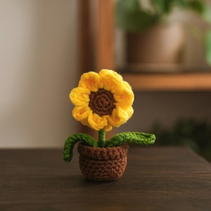 以下が含まれることがあります： 茶色の鉢に入ったかぎ針編みのヒマワリ。花は黄色の花びらと茶色の中心部を持っています。茎と葉は緑色です。鉢は小さく丸い形をしています。ヒマワリは濃い木製の表面に置かれています。