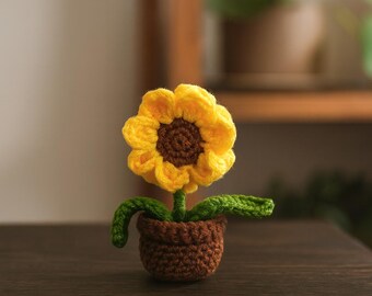 Op de afbeelding: Een gehaakte zonnebloem in een bruine pot. De bloem heeft gele bloemblaadjes en een bruin hart. De stengel en bladeren zijn groen. De pot heeft een kleine, ronde vorm. De zonnebloem staat op een donker houten oppervlak.