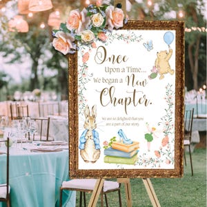 May include: A framed sign with the text "Once Upon a Time... We began a New Chapter." Features illustrations of Peter Rabbit, Winnie the Pooh, and a fairy. The sign is decorated with flowers and sits on an easel, suitable for a wedding or event.