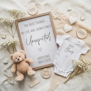 Może przedstawiać: Ogłoszenie o narodzinach dziecka z białym body z napisem "Baby Oliver", tablicą literową z napisem "Najlepsze rzeczy w życiu są czasem nieoczekiwane, przybycie maj 2026" i misiem. Drewniane akcenty i kwiaty dopełniają scenę.