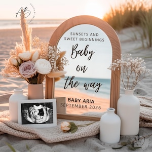 May include: A pregnancy announcement featuring a mirror with the words "Baby on the way." The image includes flowers, an ultrasound scan, and white vases, arranged on a sandy beach setting.