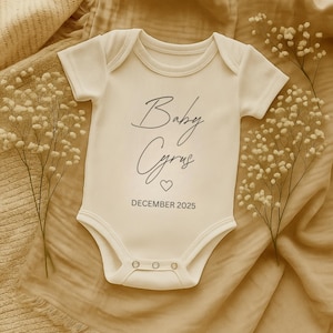 Könnte beinhalten: Cremefarbener Baby-Body mit dem Text "Baby Cyrus" in Schreibschrift, einem Herzsymbol und "DECEMBER 2025". Der Body hat kurze Ärmel und drei Druckknöpfe. Der Hintergrund ist ein beiger Stoff, mit weißen Blütenzweigen.