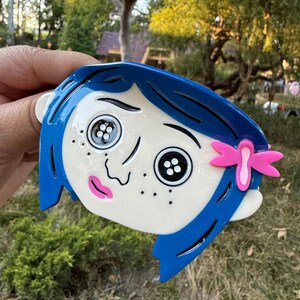 May include: A blue and white decorative item featuring a cartoon character's face with button eyes, blue hair, and a pink bow. The character has freckles and a pink mouth. The item is held against a background of green grass and trees.
