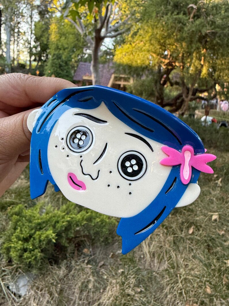 May include: A blue and white decorative item featuring a cartoon character's face with button eyes, blue hair, and a pink bow. The character has freckles and a pink mouth. The item is held against a background of green grass and trees.