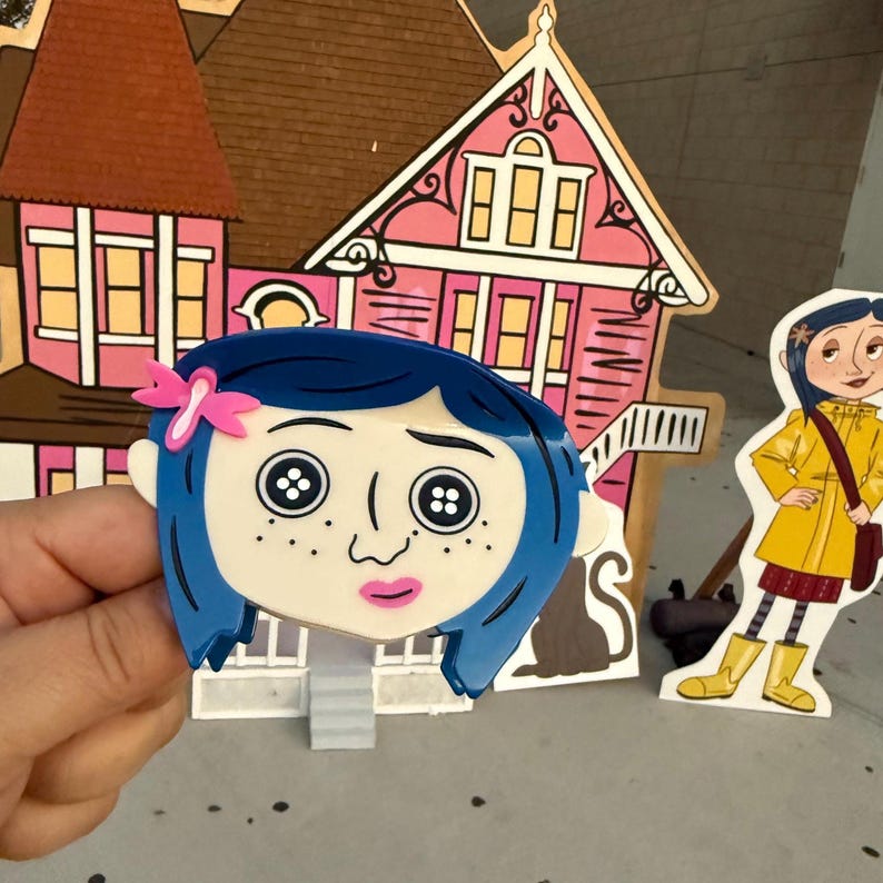 May include: A blue-haired character face with button eyes and a pink bow, held in front of a pink and brown house cutout. A yellow-coated character cutout stands nearby. The scene is reminiscent of a children's story.