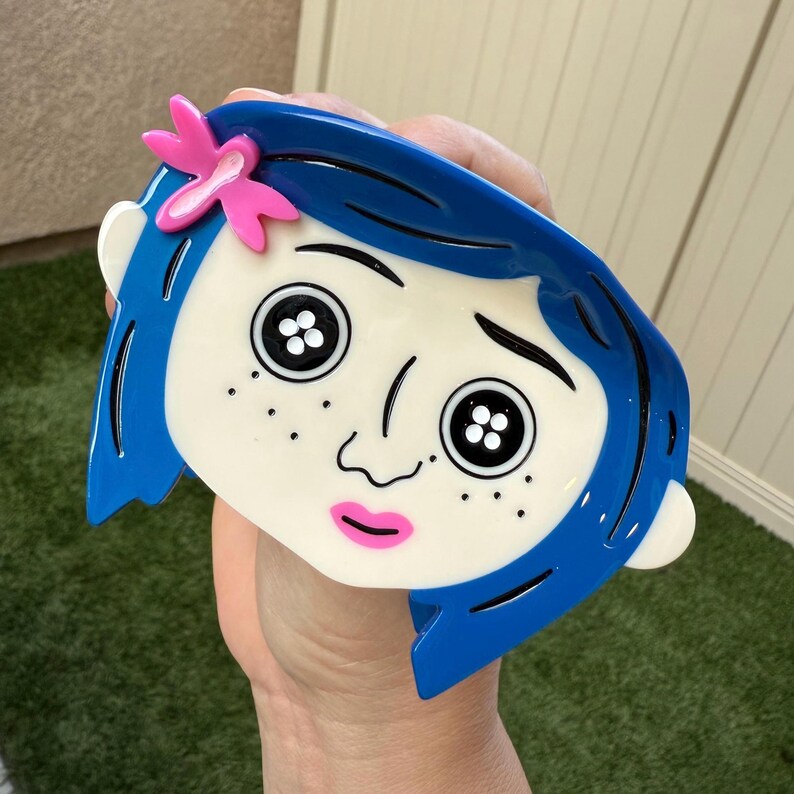 May include: A blue and white face-shaped accessory with black button eyes, freckles, and a pink bow. The accessory has blue hair and a pink mouth. The design is a cartoon character.
