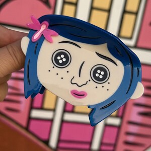 May include: A blue and white cartoon character brooch with a pink bow. The brooch features a character with blue hair, button eyes, and freckles. The background is a pink and white illustration of a house.