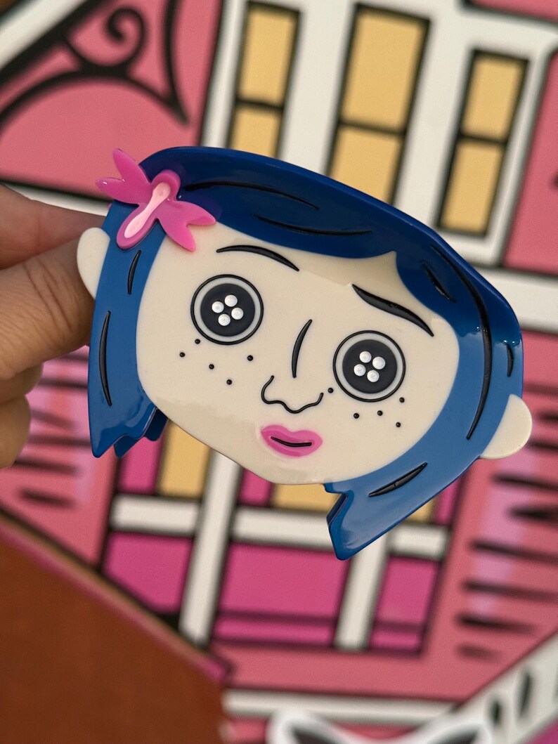 May include: A blue and white cartoon character brooch with a pink bow. The brooch features a character with blue hair, button eyes, and freckles. The background is a pink and white illustration of a house.